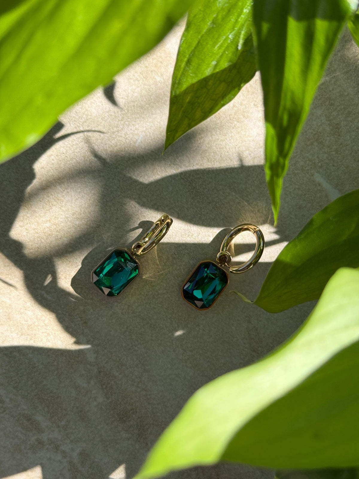 Taimana Kākāriki Gold Huggies with green stones, among leaves