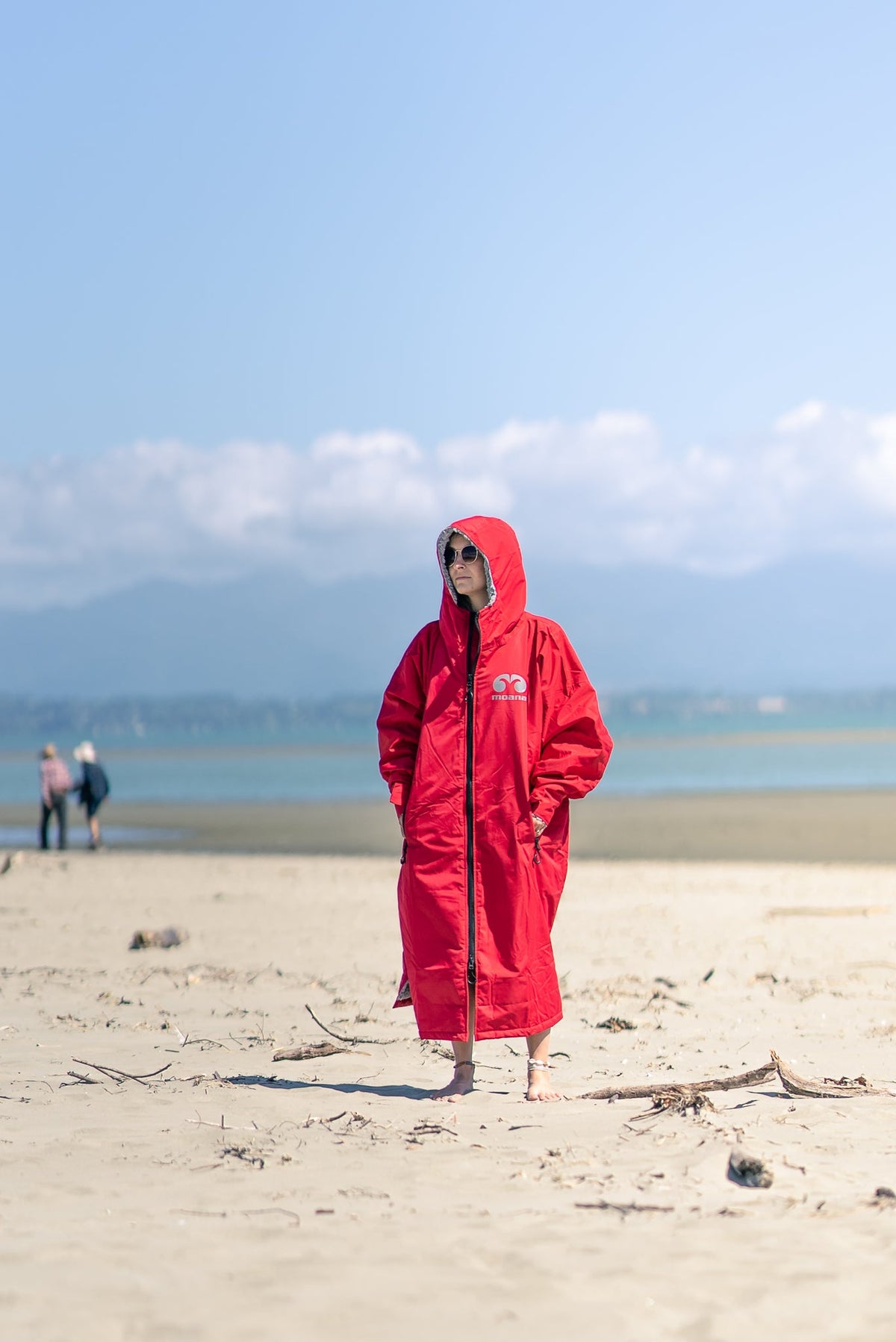 Person standing on a beach in a red Moana SUP and Surf change robe, hood up