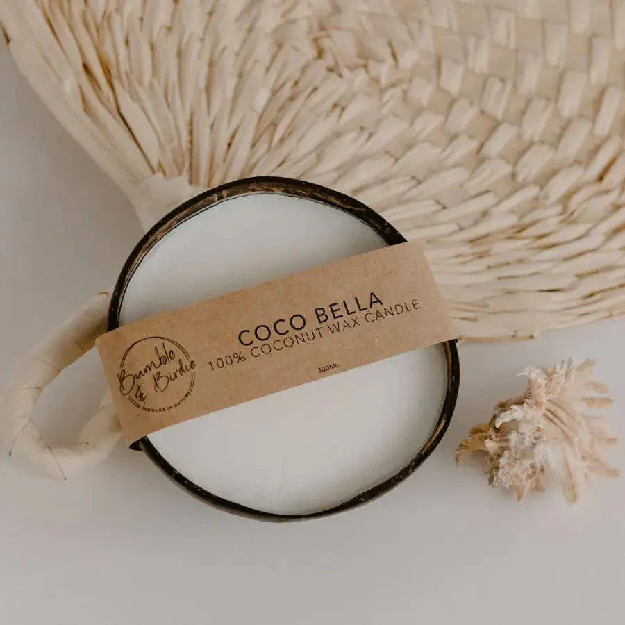 Coco Bella pure coconut candle in a coconut shell with a Bumble & Birdie label, beside a woven fan and shells