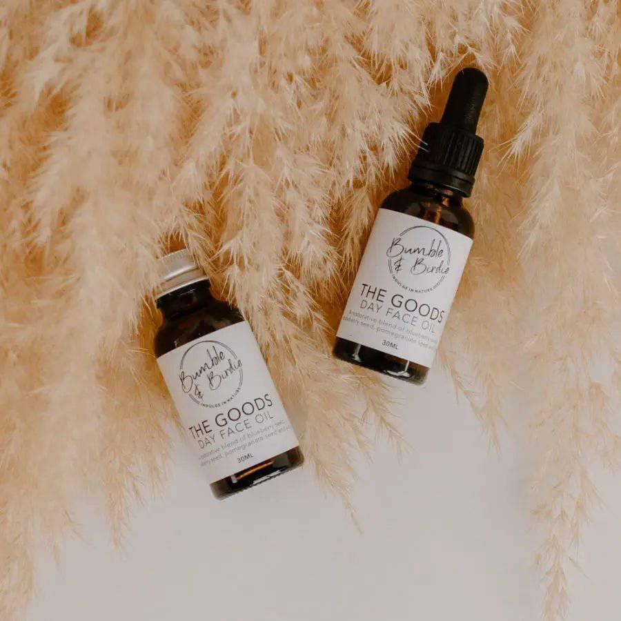 Two bottles of The Goods day face oil lying on pampas grass
