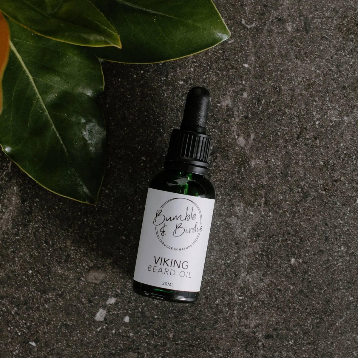 Viking beard oil in a dropper bottle by Bumble & Birdie, on dark stone with leaves