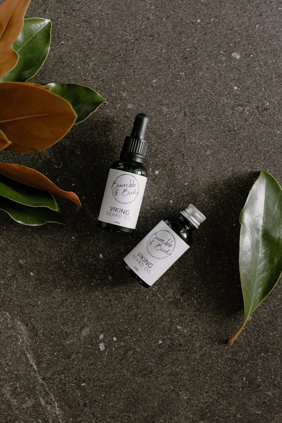 Two Viking beard oil bottles on dark stone with magnolia leaves