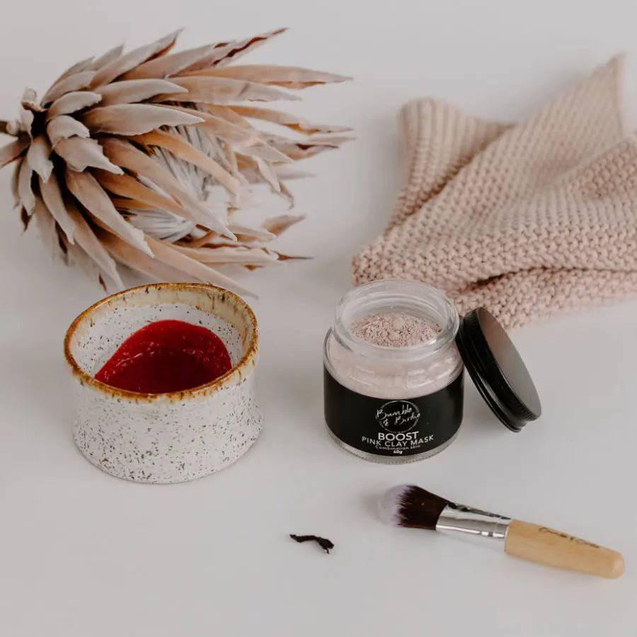 Boost Pink Clay Mask jar by Bumble & Birdie beside a bowl of mixed pink mask, a brush and dried protea
