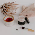 Boost Pink Clay Mask jar by Bumble & Birdie beside a bowl of mixed pink mask, a brush and dried protea