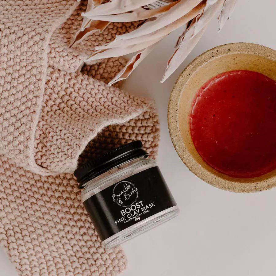 Boost Pink Clay Mask jar on a knitted cloth beside a bowl of mixed pink mask