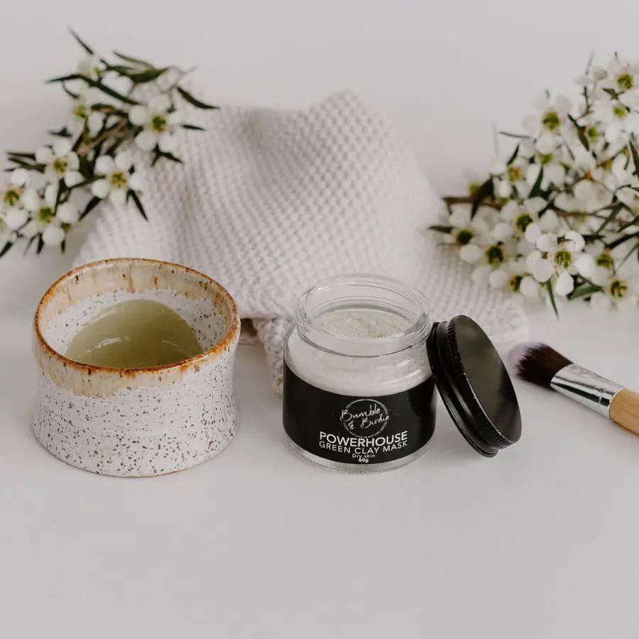 Powerhouse Green Clay Mask jar with a bowl of mixed green mask, a brush and white flowers
