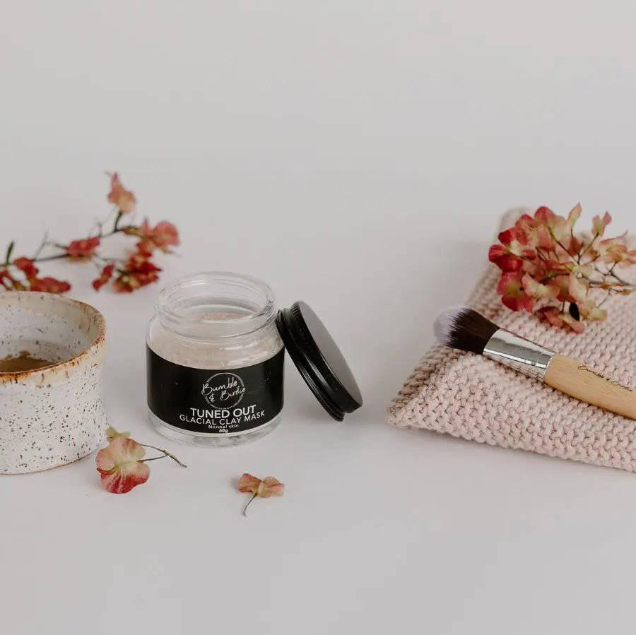 Tuned Out Glacial Clay Mask jar with a speckled clay bowl, mask brush and dried flowers