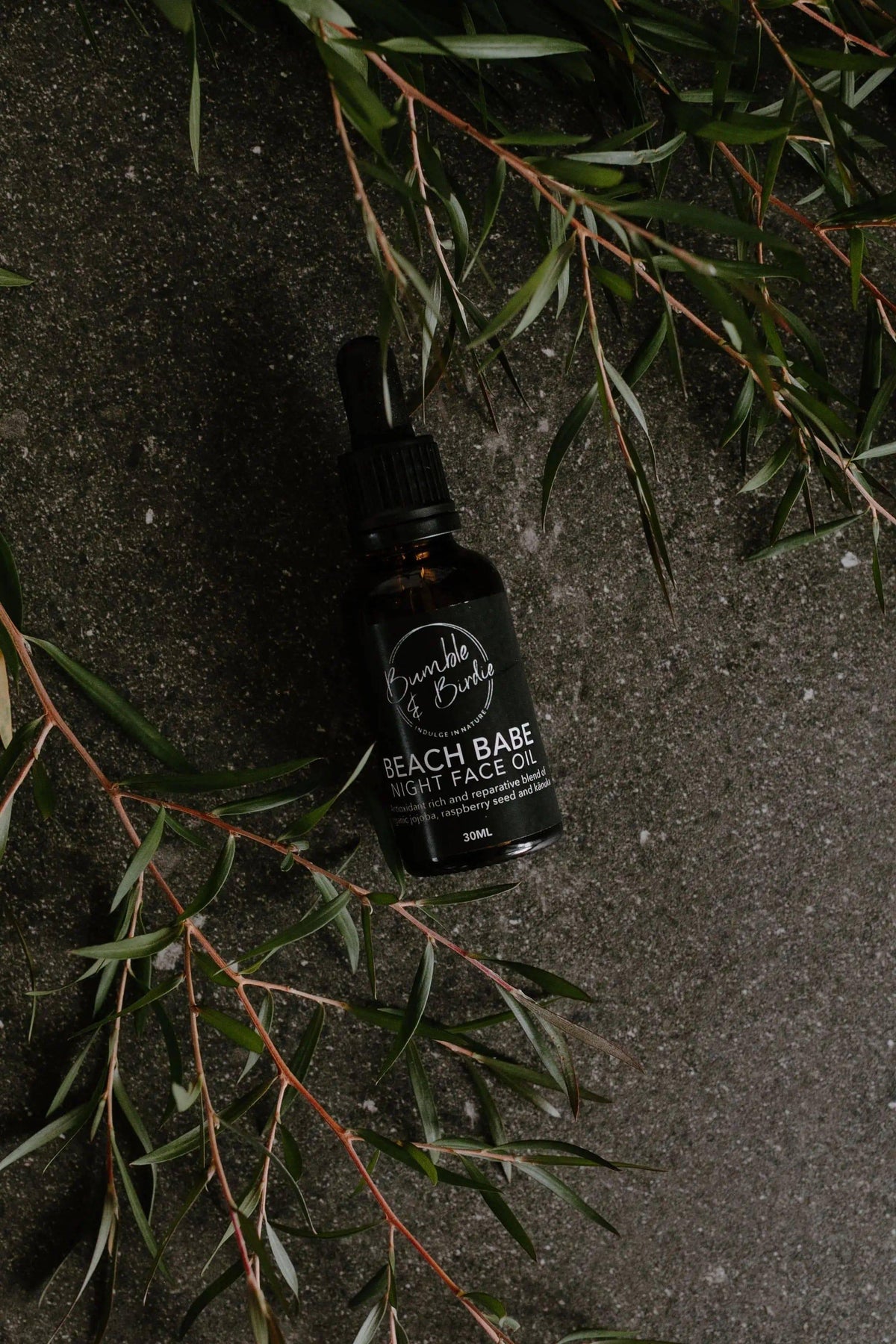 Beach Babe night face oil bottle on dark stone with green sprigs