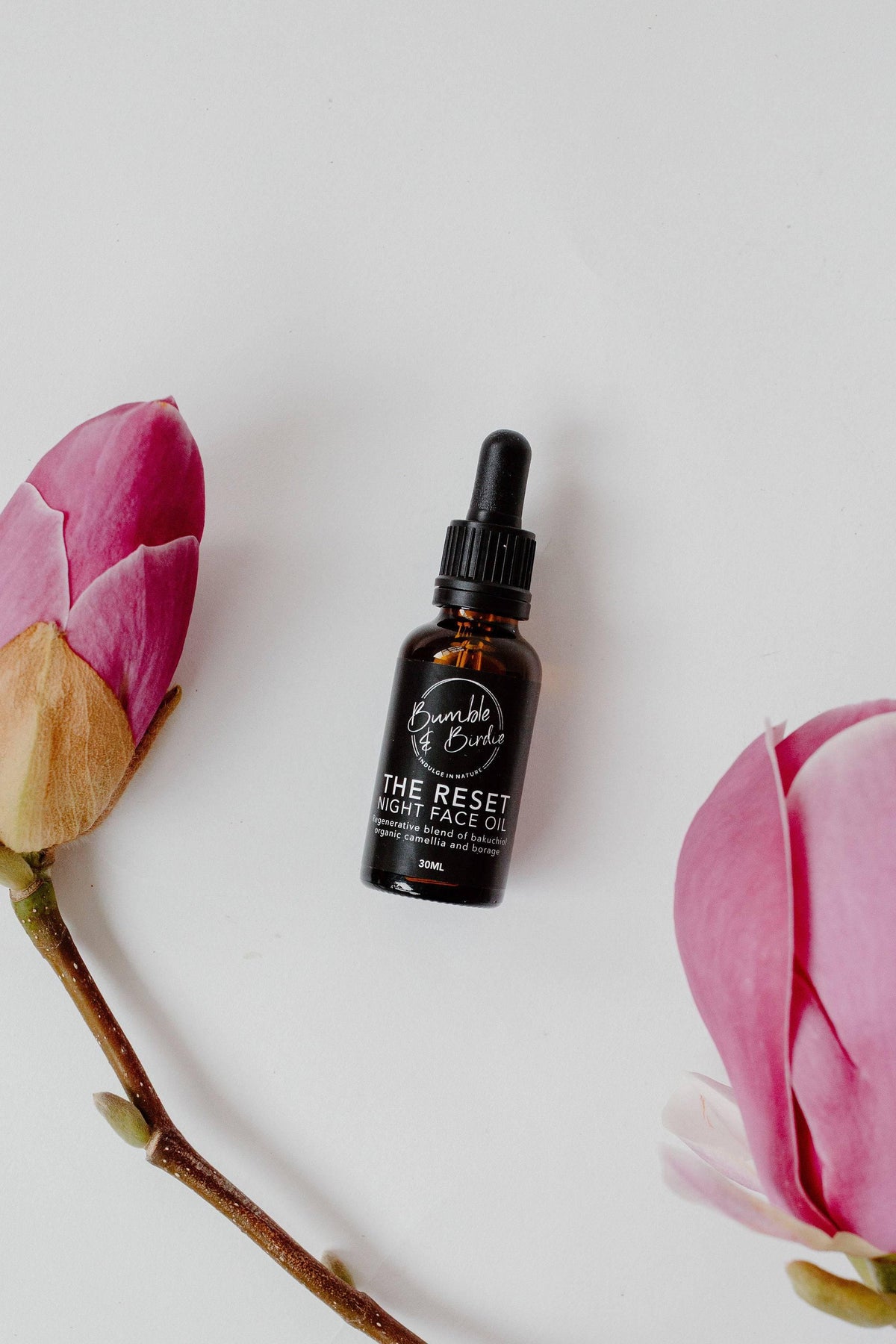 The Reset night face oil dropper bottle between pink magnolia flowers