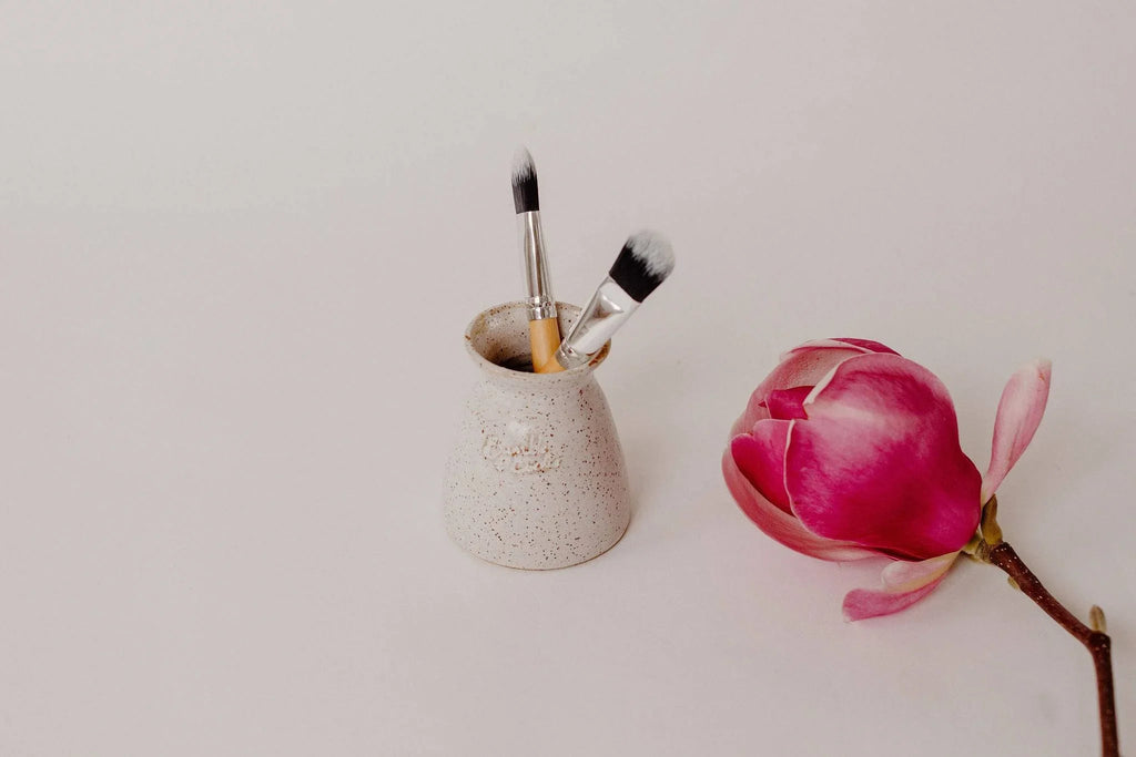Earth Brush Haven speckled pottery vase holding face mask brushes, beside a magnolia flower
