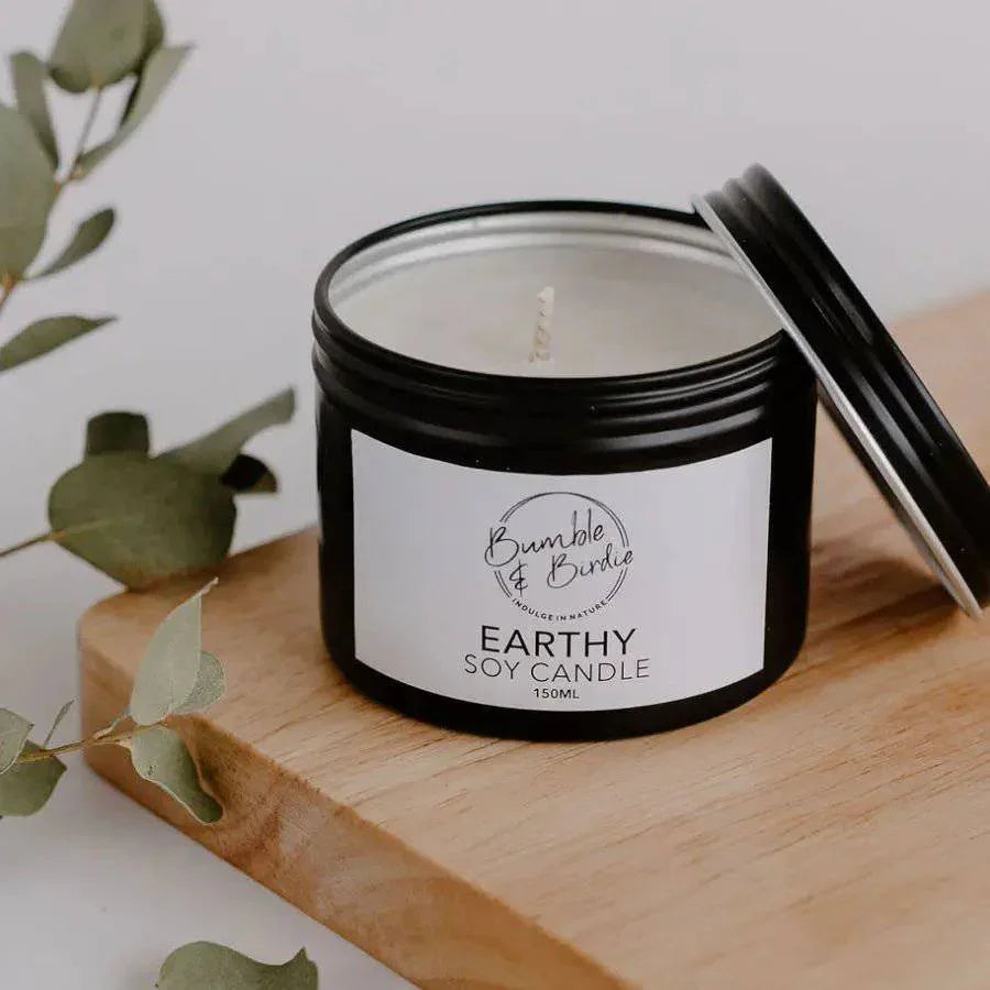 Earthy soy candle in an open black tin by Bumble & Birdie, on a wooden board with eucalyptus