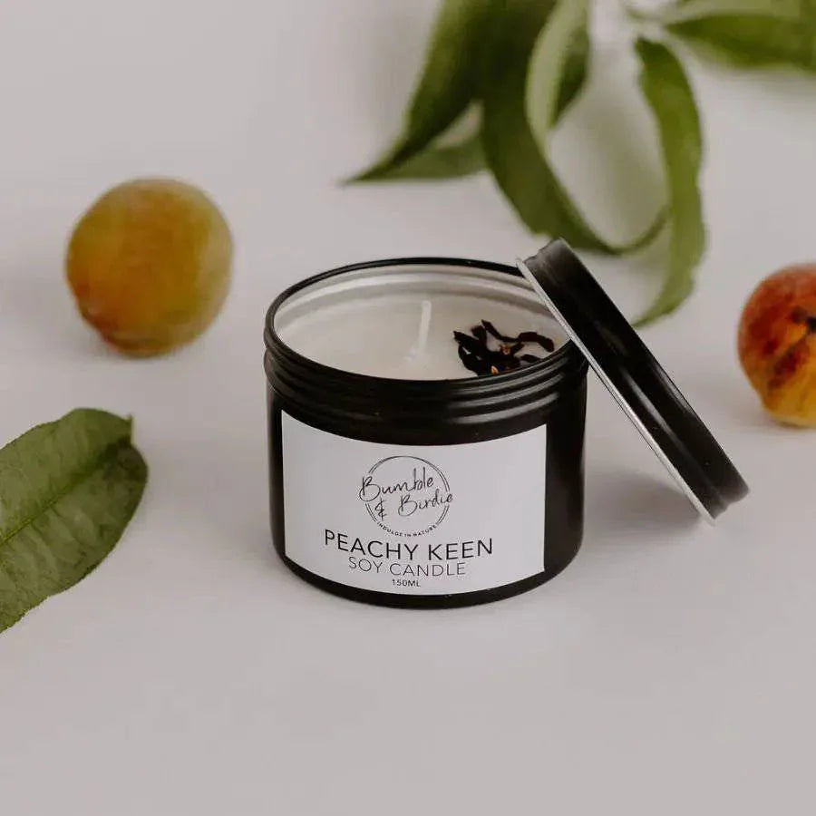 Peachy Keen soy tin candle with its lid off, beside peaches and leaves